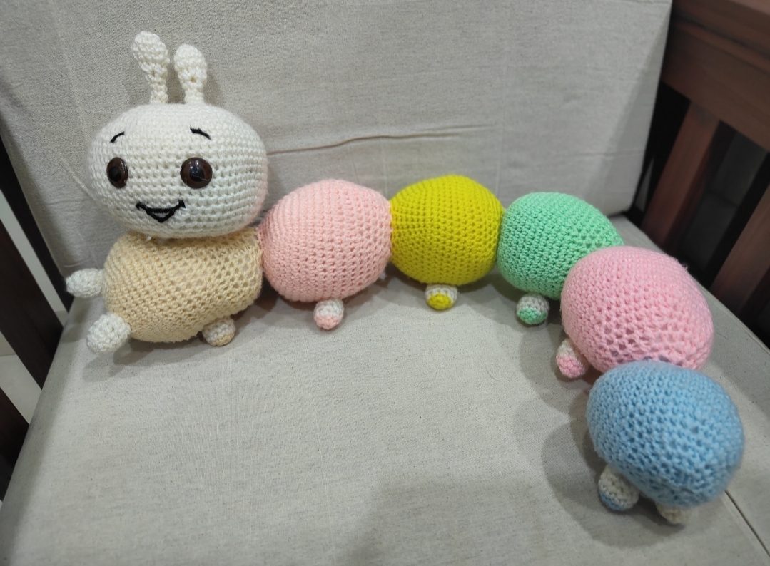 The Pastel Caterpillar — a long crocheted caterpillar with a smiling cream face and pastel body segments in peach, pink, yellow, mint, pink and blue